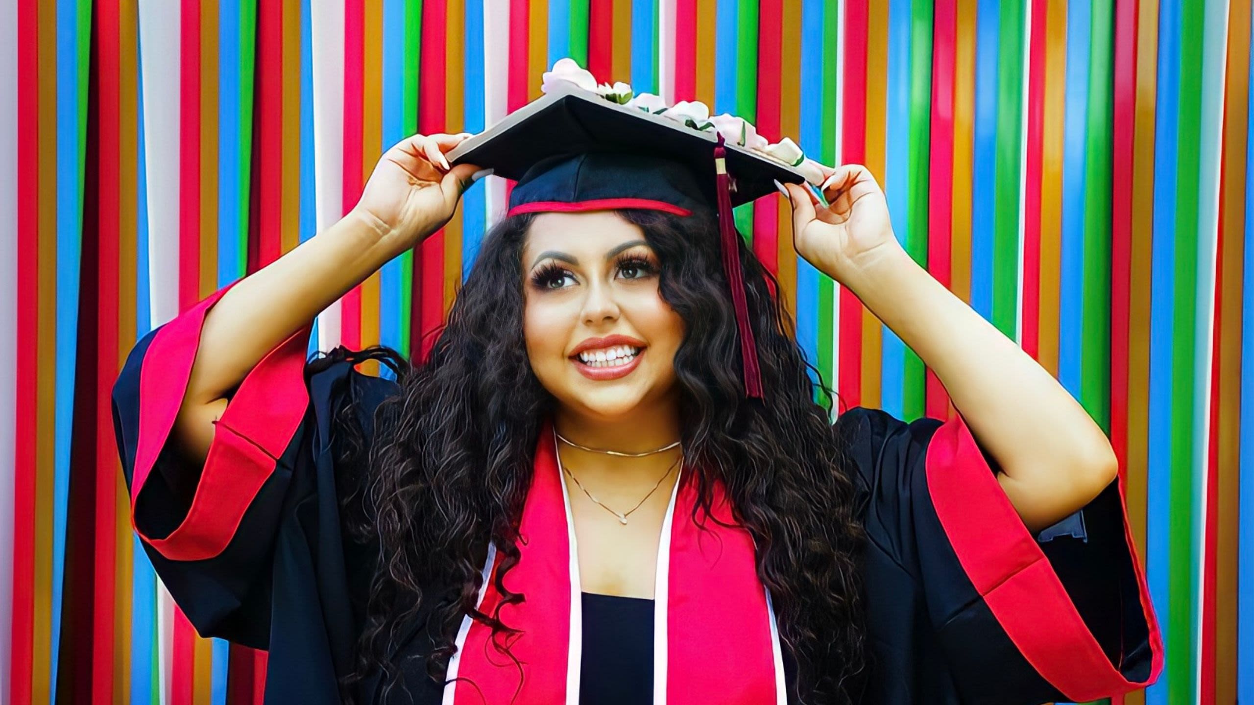 Graduate in regalia smiling in front of striped art wall holding grad cap