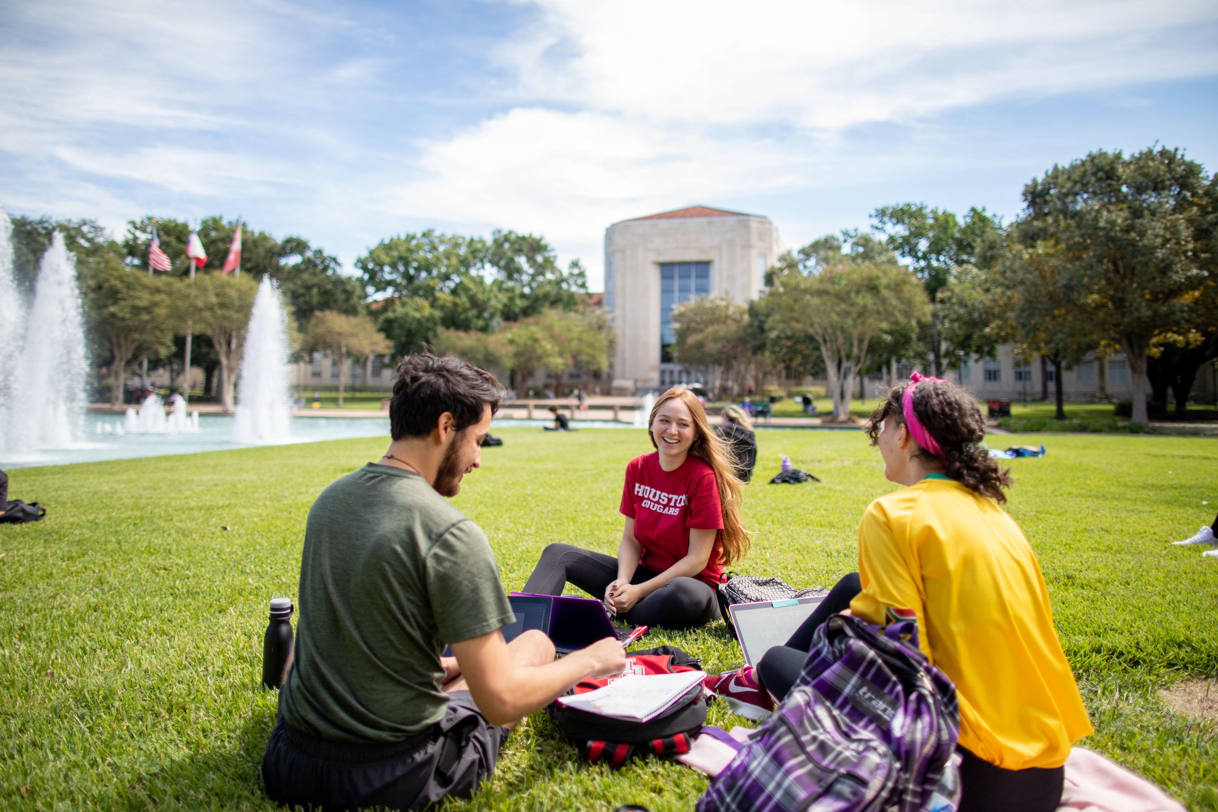 Three students study together on laptops while sitting in the grass of the Cullen Family Plaza. They are surrounded by the iconic fountains.