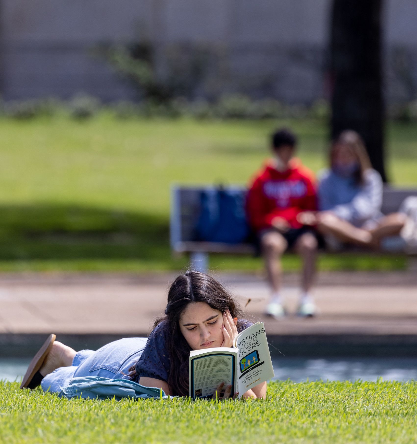 A student reading a book and lying on the grass while two students sit on a bench in background