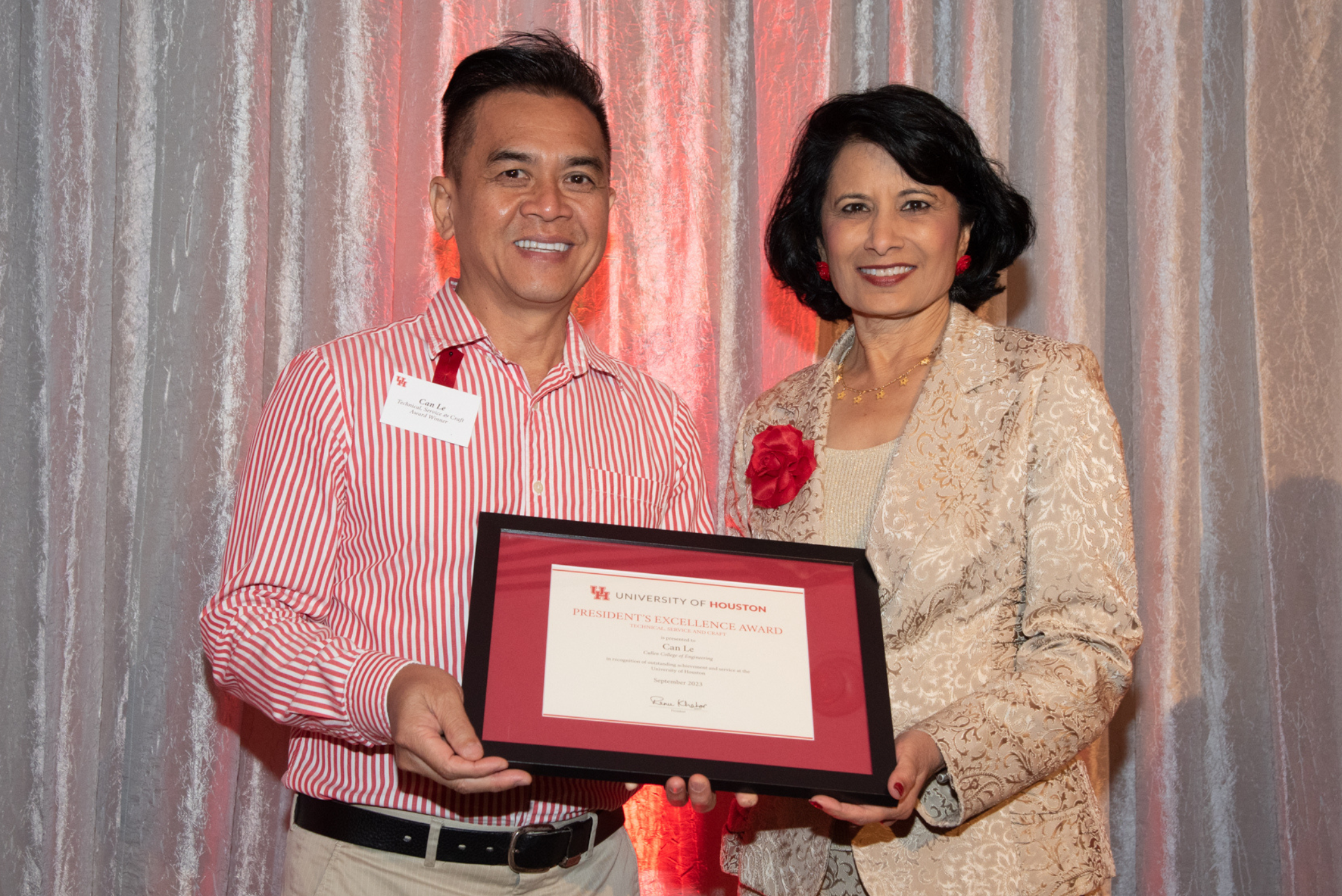 Standing in front of a silver curtain holding a framed certificat are President Renu Khator in a gold suit and Technical, Service and Craft Award winner, Can Le in a red and white striped dress shirt.