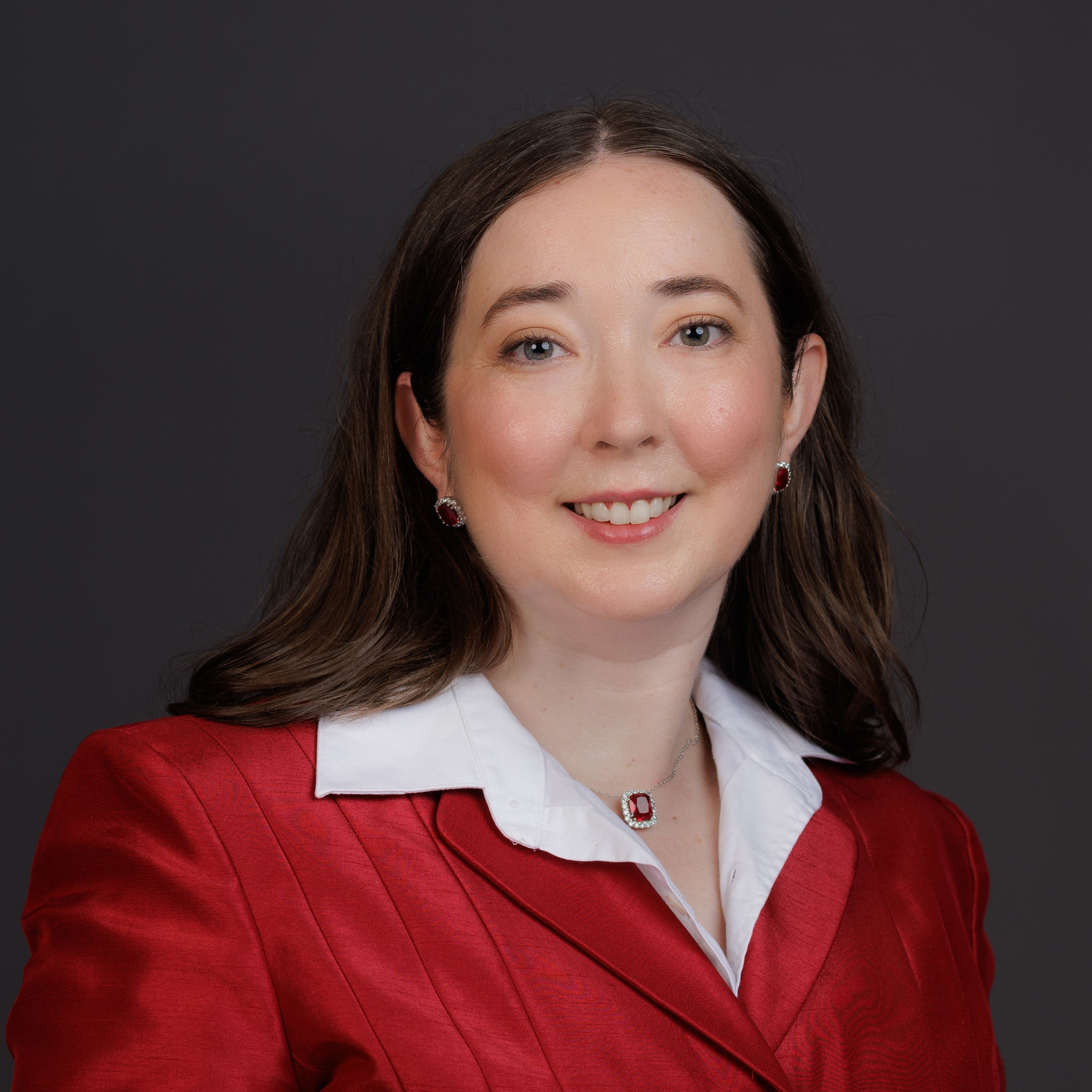 Portrait of Holley Love wearing a red suit and red jewelry, smiling in front of a gray background.