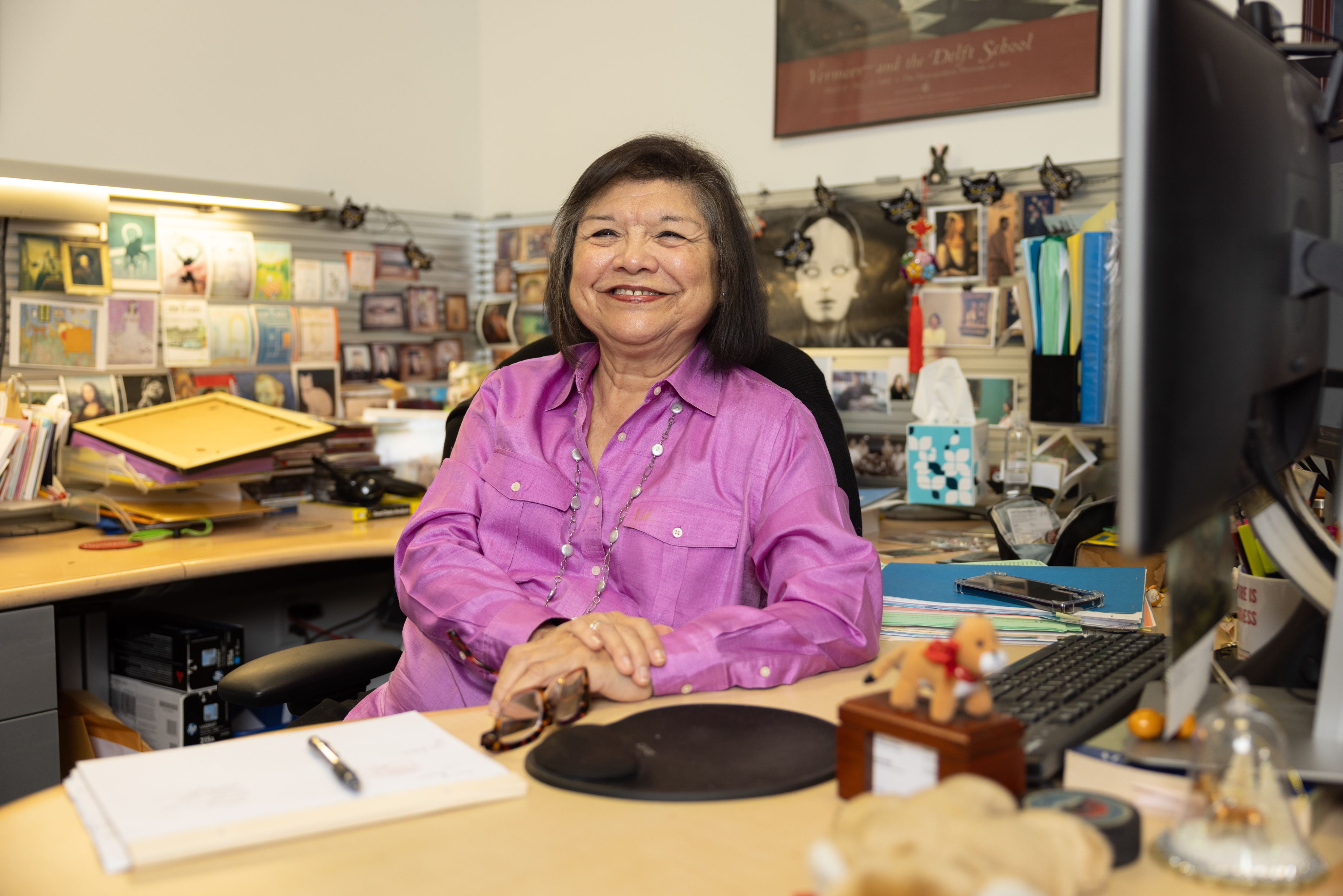 Mary Gould in a pretty purple shirt sitting in her office; in the background is a collection of her memorabilia. 