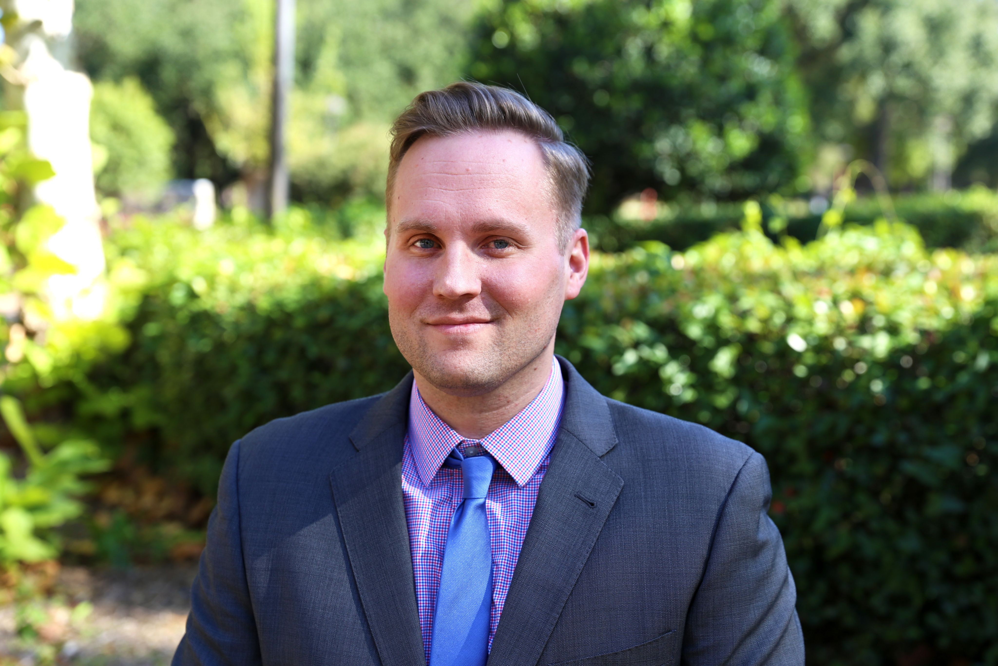 Scott Radimer smiling in front of greenery. He is wearing a grey suit with a blue tie.