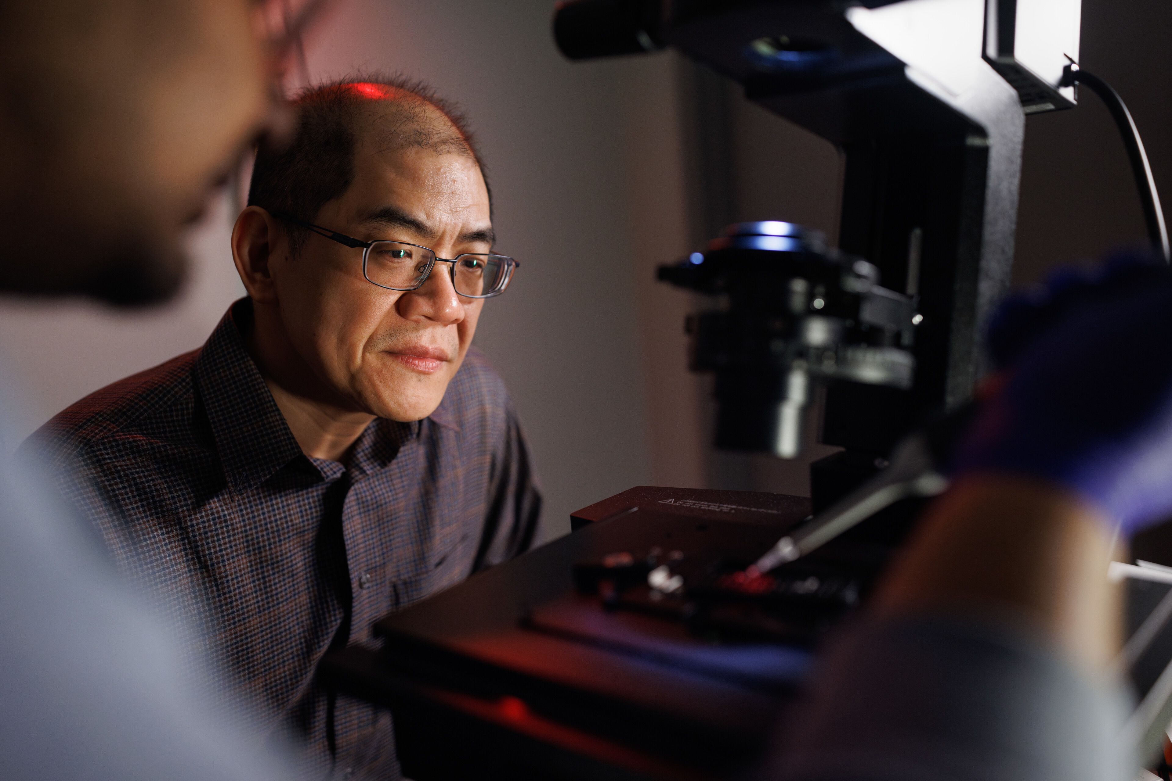 A man examines a microscope intently, while another man observes him with interest in a laboratory setting.