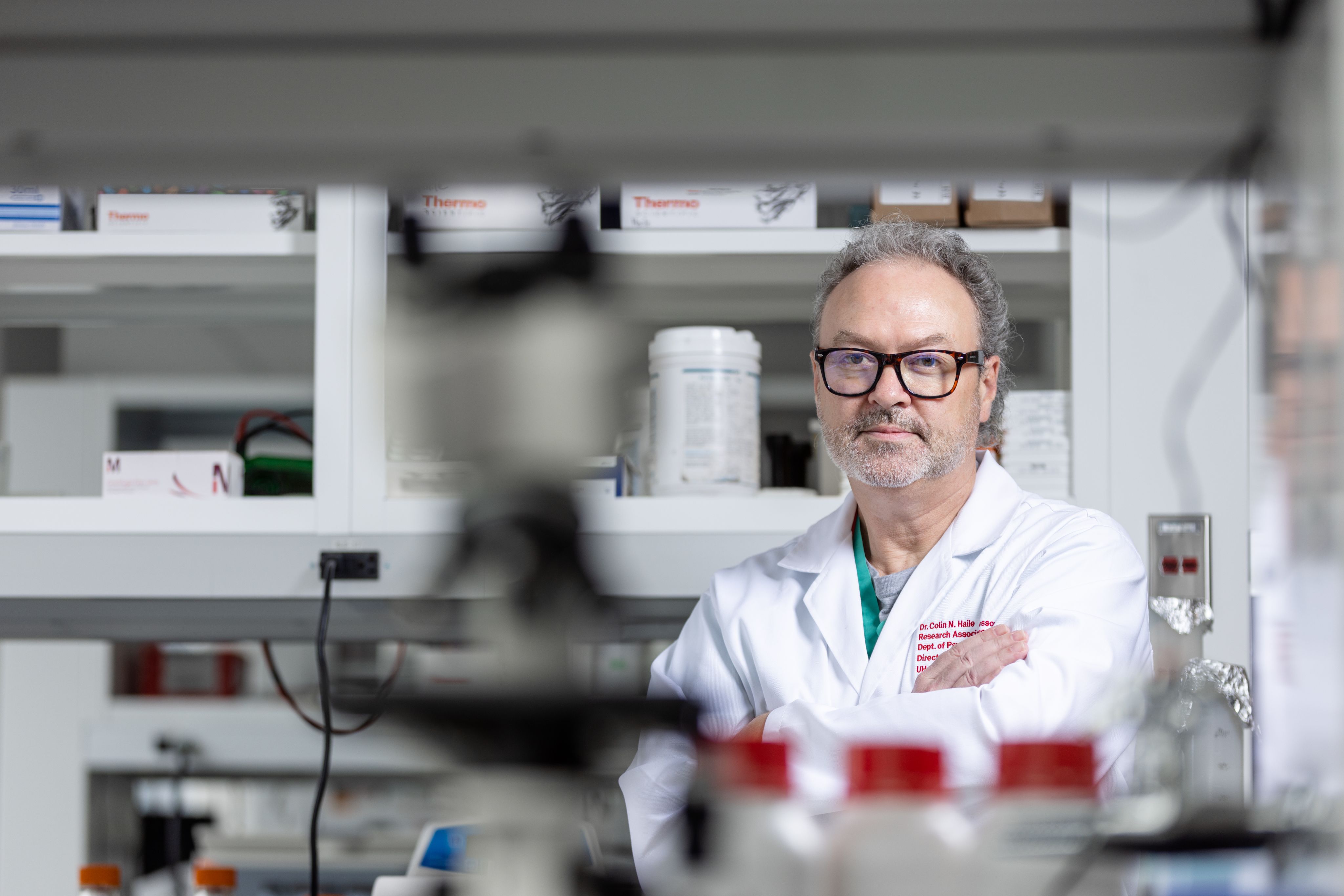 Colin Haile in a lab coat stands confidently in front of a shelf filled with various scientific equipment and materials.