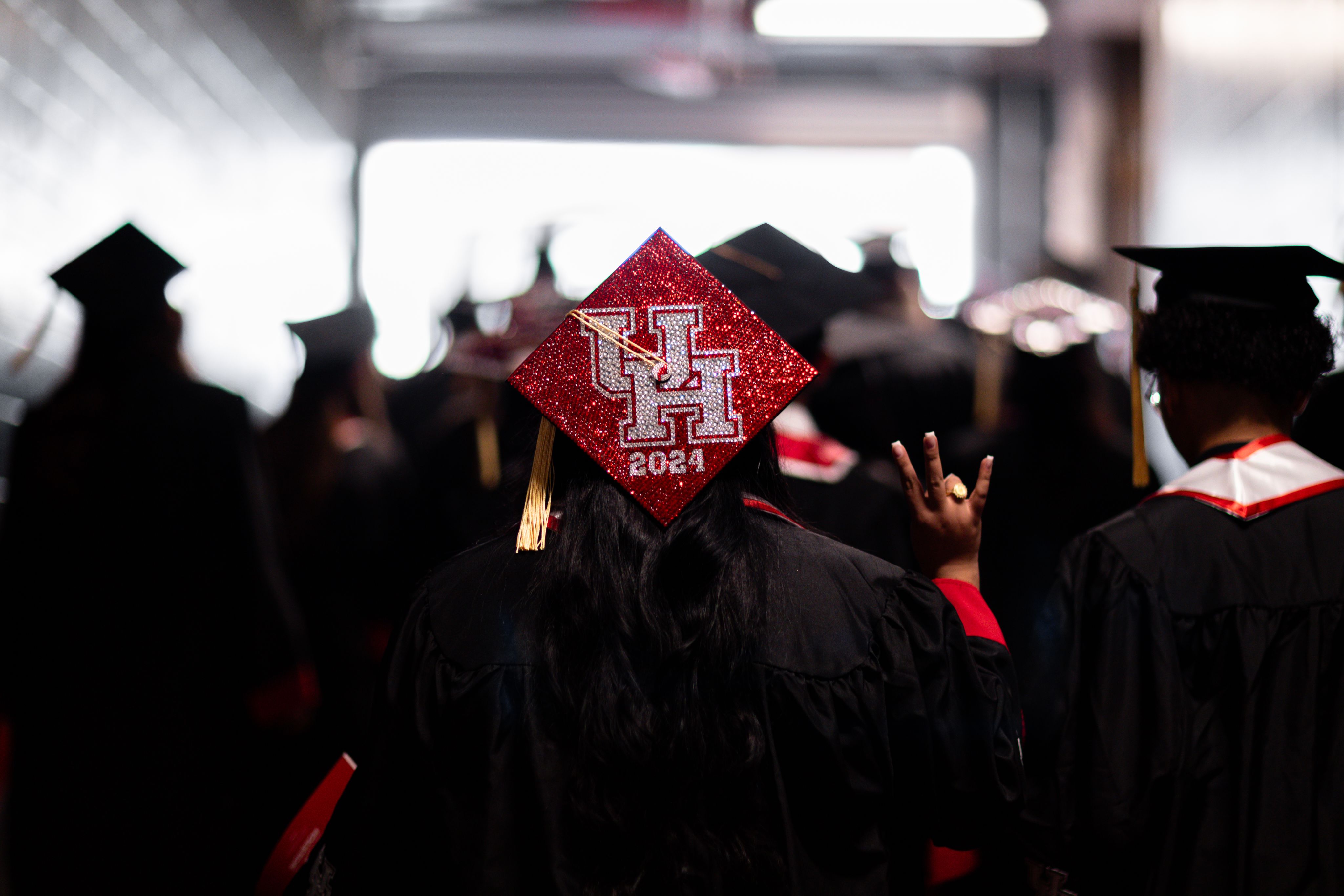 Female student with back to the camera, wearing red sparkled graducation cap with UH 2024 emblazoned on it and holding up the Cougar paw sign.