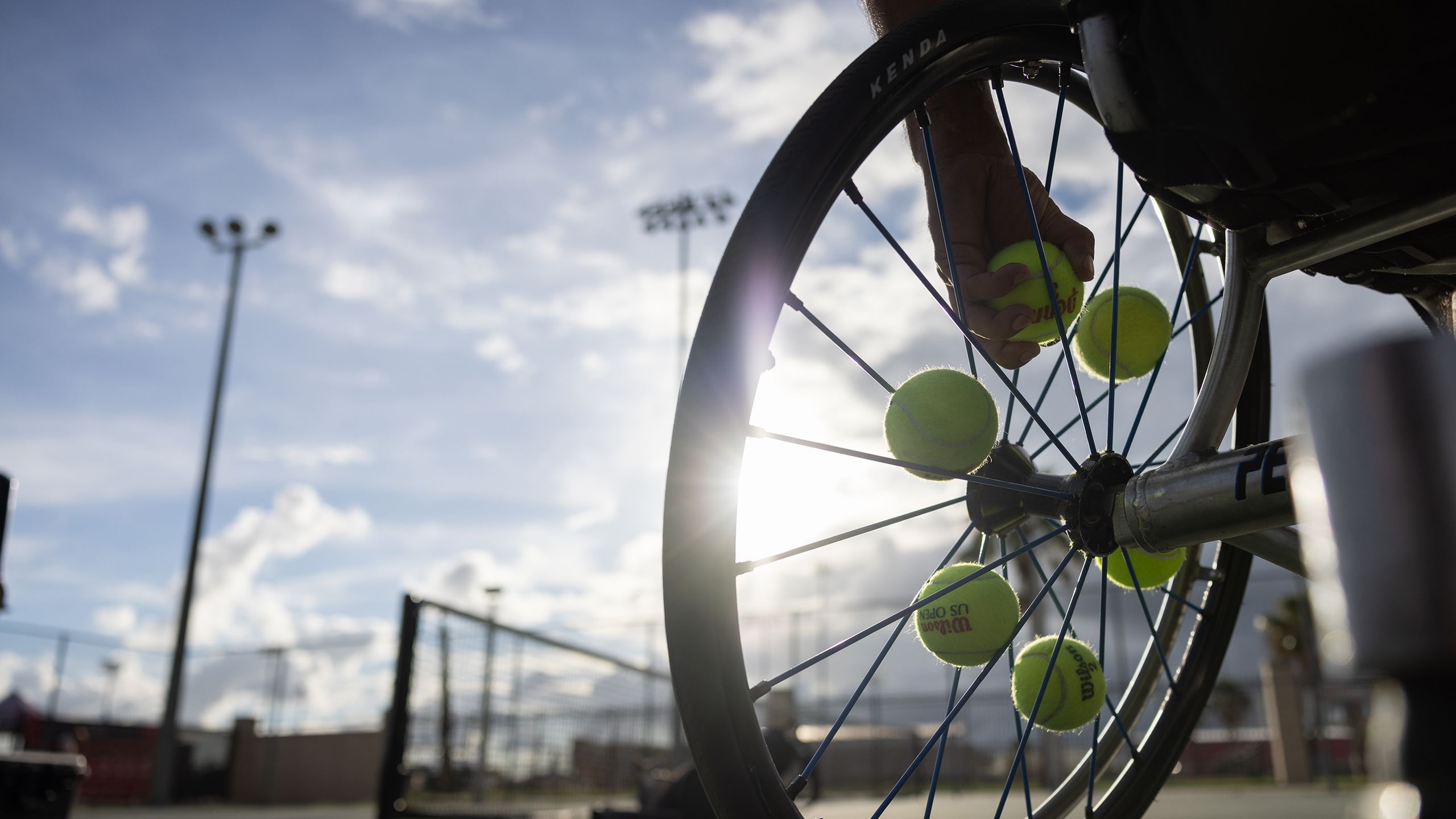 Image of a wheelchair wheel with tennis balls in the spokes and a person's hand grasping a tennis ball.