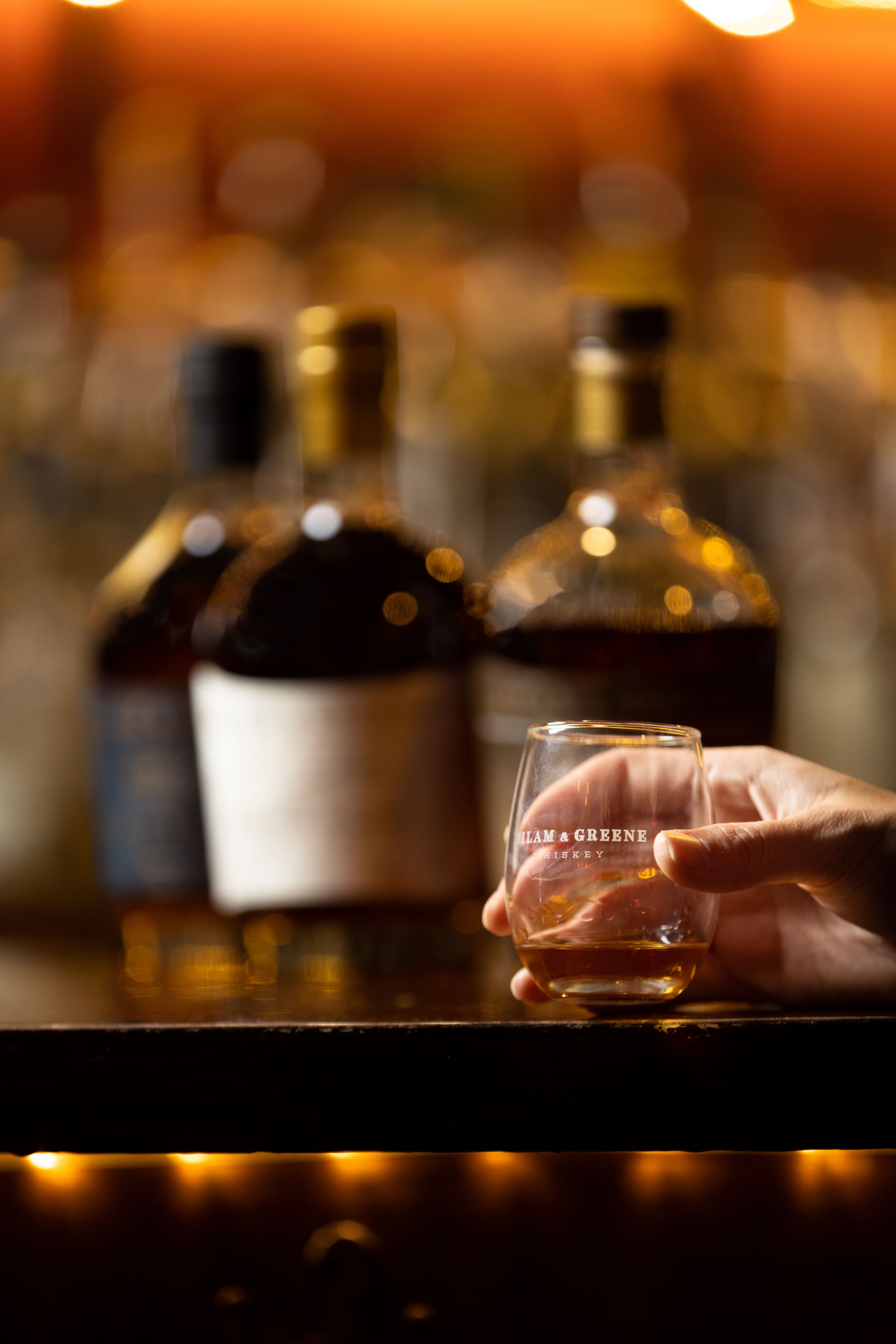 A woman's hand holding a shot of whisky, in front of 3 bottles of whisky.