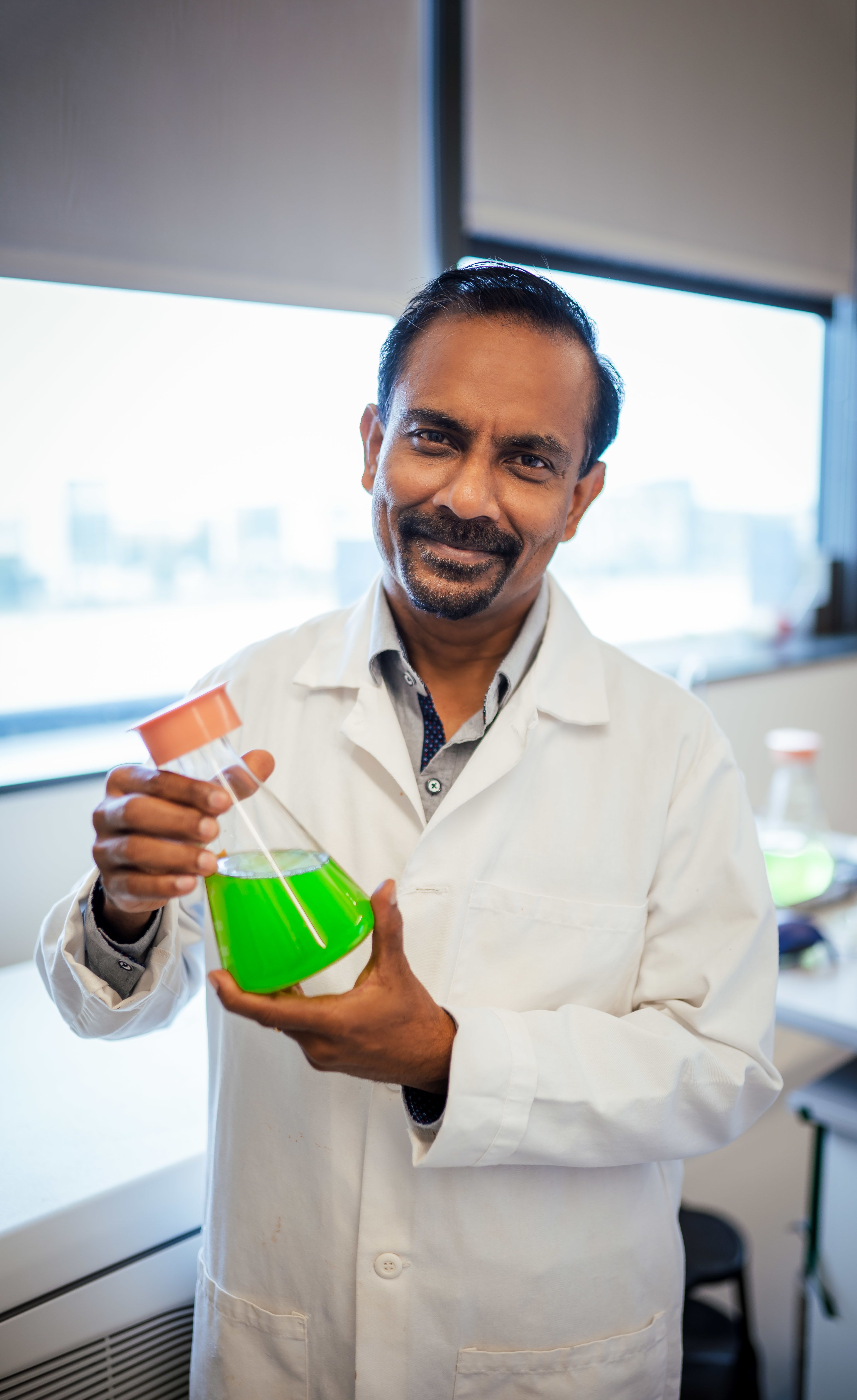 Venkatesh Balan wearing a white lab coat & holding a beaker half full of neon green liquid