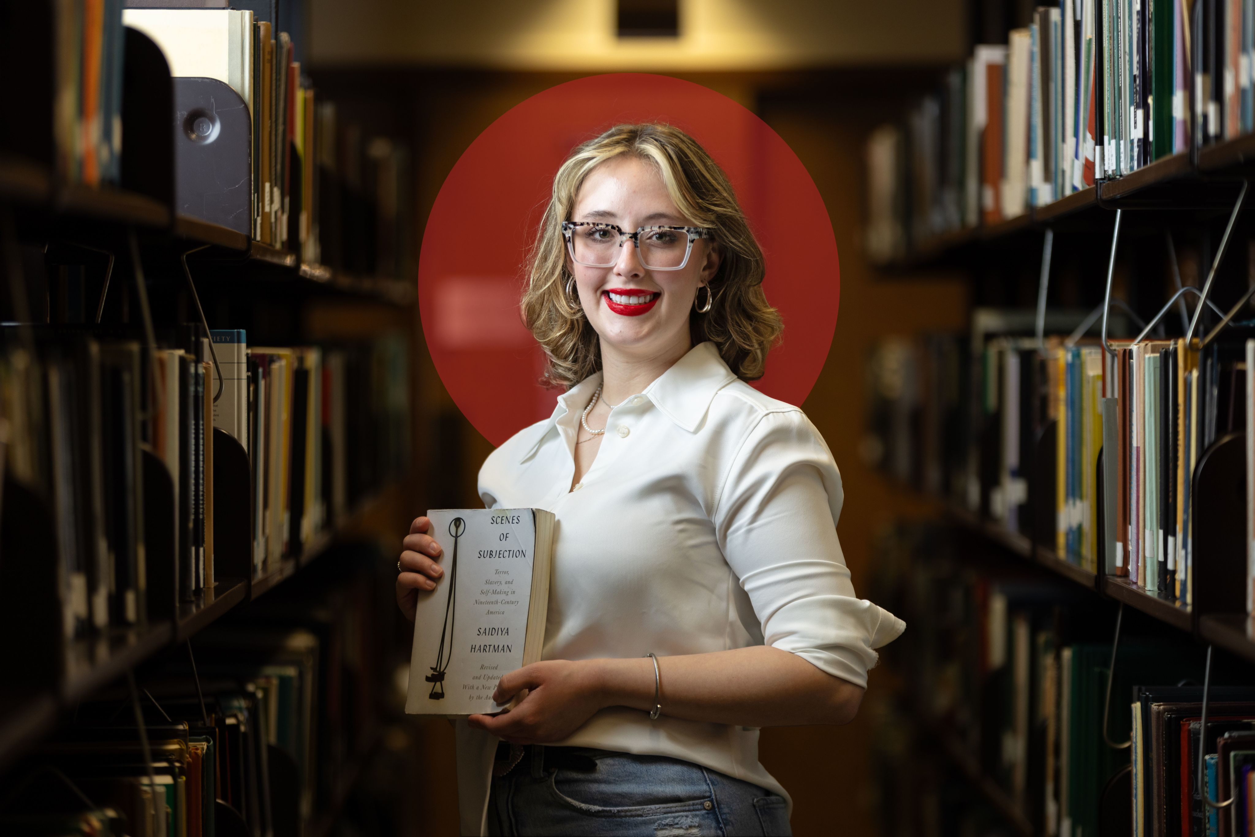 Portrait of Alissa Boxleitner holding a copy of "Scenes of Subjection" between two rows of library shelves.
