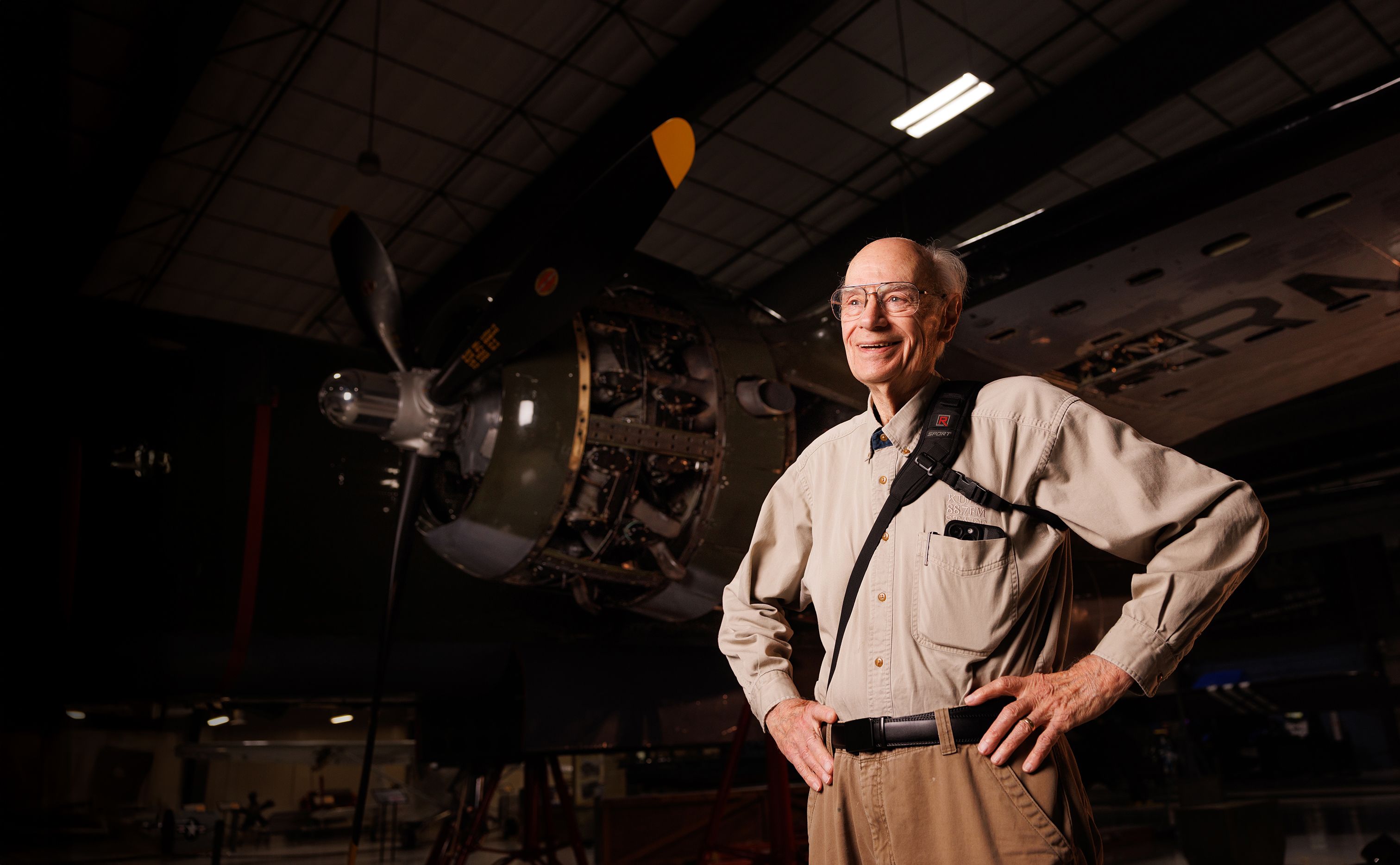 Portrait of John Lienhard, who stands smiling with his hands on his hips. He stands in front of a large engine and wears a camera strap slung around his body. 