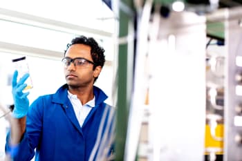 This is a photo of Praveen Bollini, associate professor of chemical engineering at UH in the lab. He is holding up a sample of liquid in a glass container and looking at it.