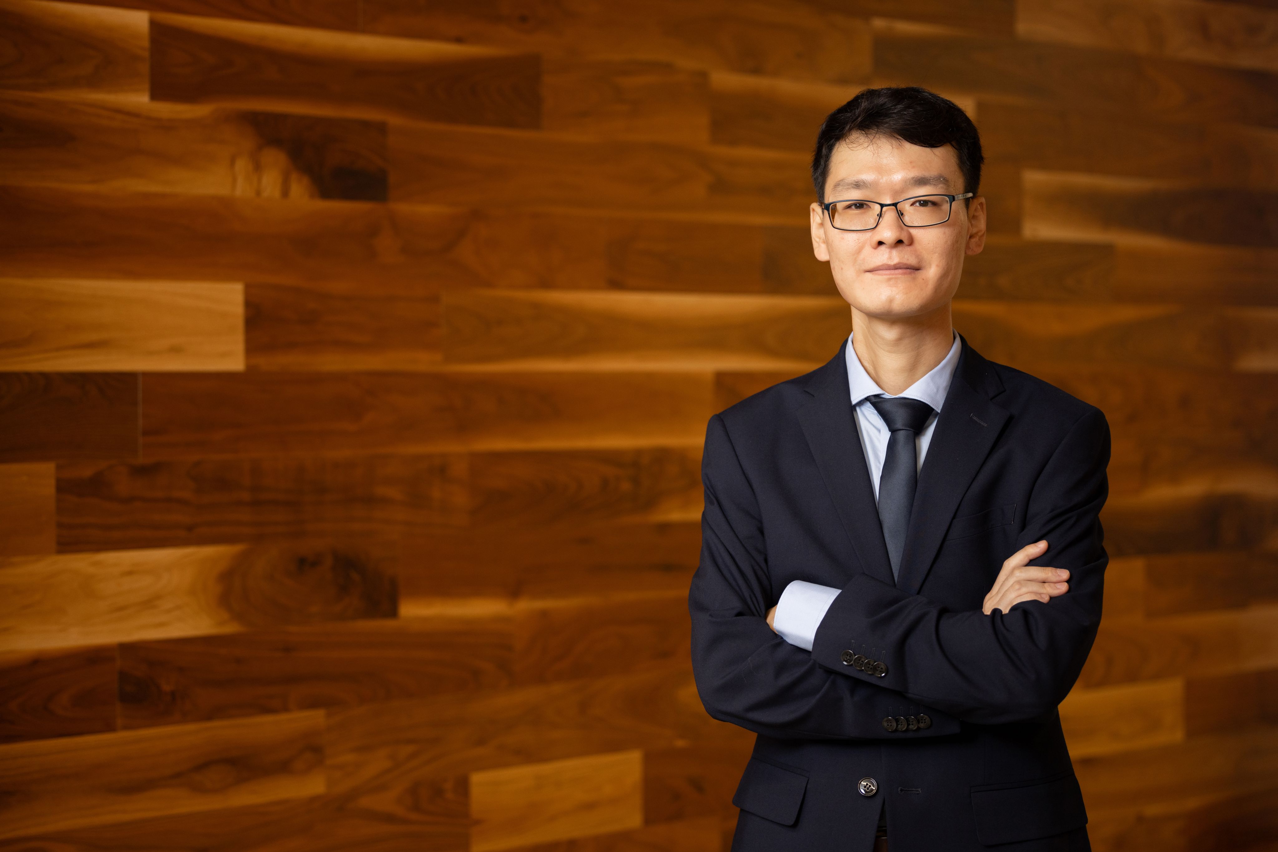 Male with dark hair, glasses wearing a dark suite with a tie standing with arms crossed in front of a wooden wall