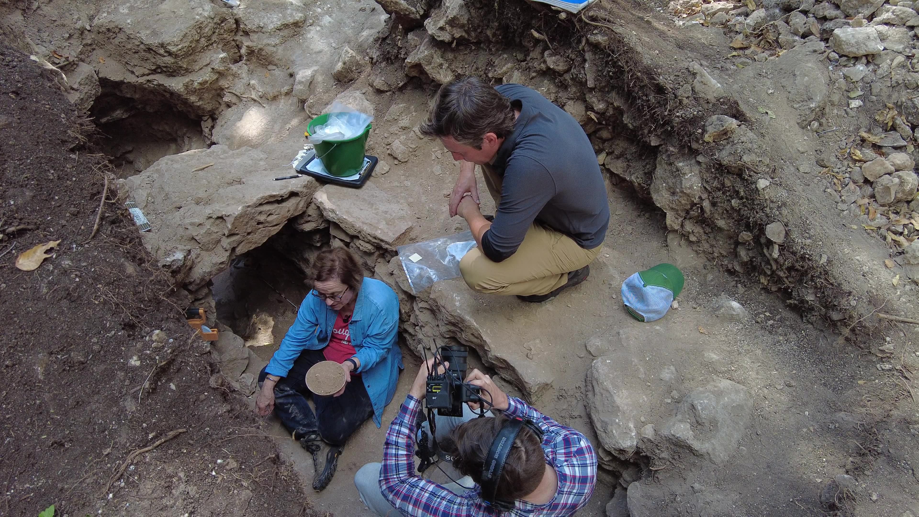 Women in blue jacket sitting in aracheological site with news crew.